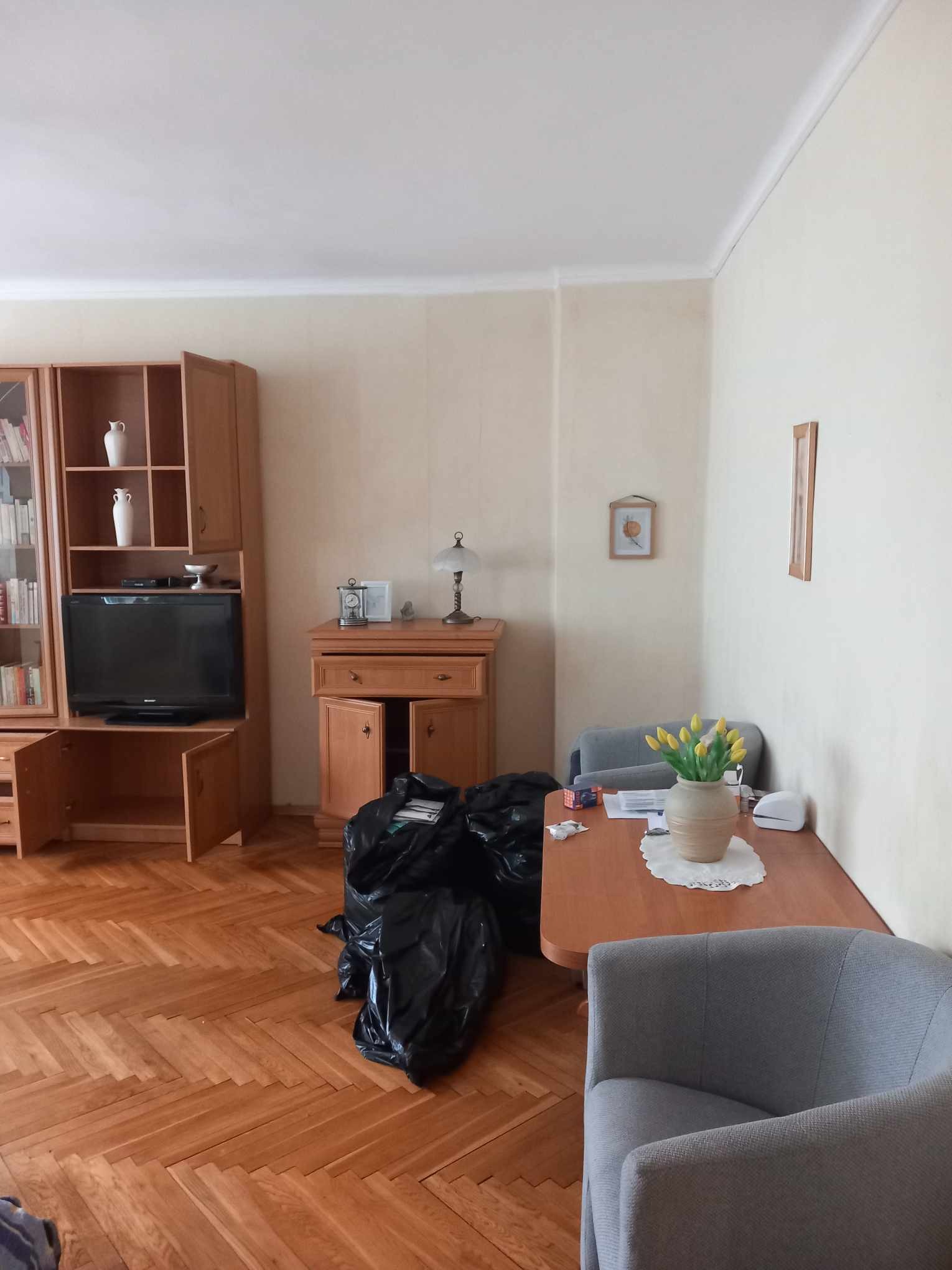 Salon przed remontem: widoczne meble w stylu retro, parkiet w jodełkę, ściany w odcieniach beżu oraz worki na śmieci sugerujące przygotowania do prac remontowych.