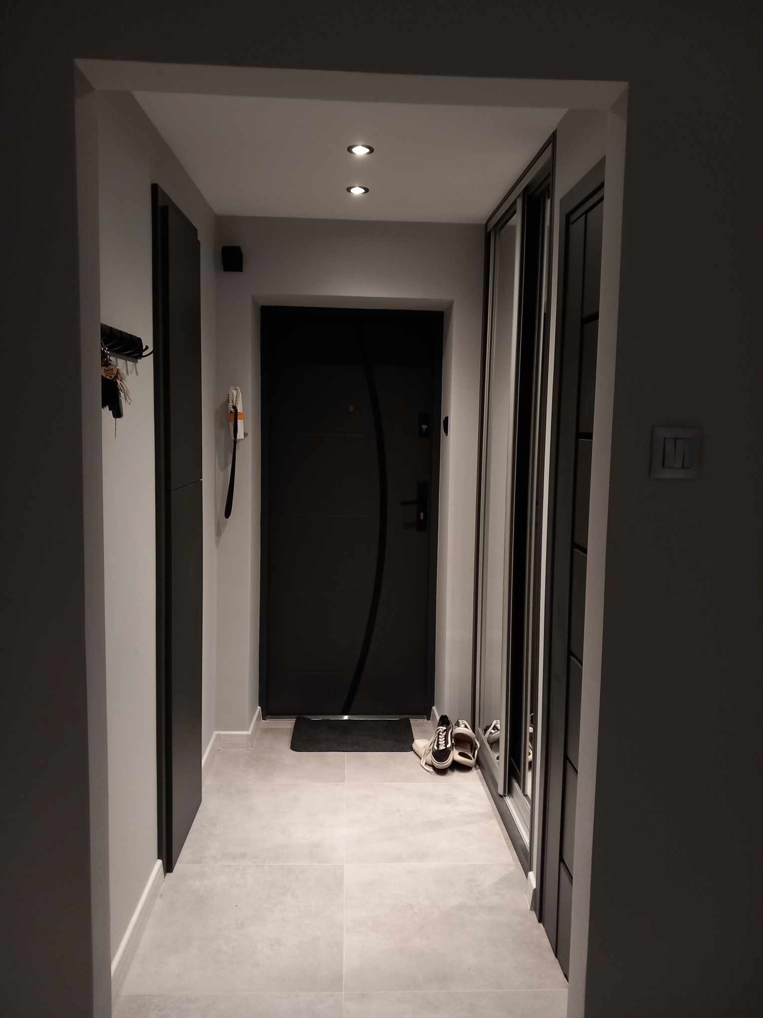 Nowoczesny przedpokój w szarościach z przesuwną szafą, minimalistycznym oświetleniem punktowym i ciemnymi drzwiami wejściowymi, na podłodze jasne płytki.