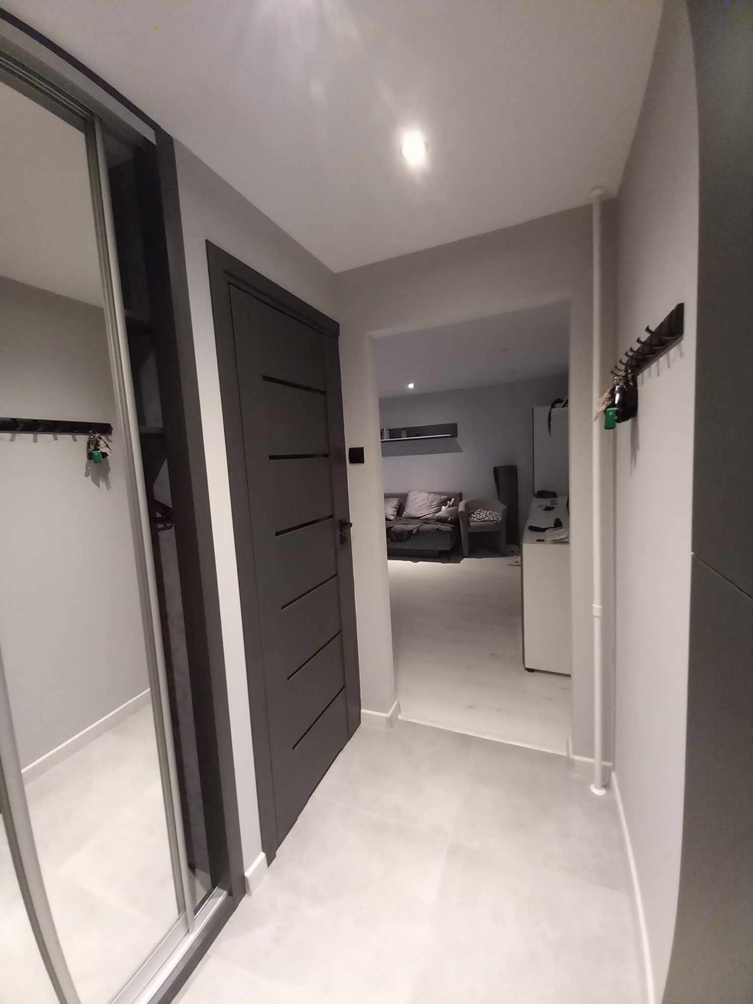 Nowoczesny przedpokój z szarą zabudową szafy przesuwnej z lustrem, ciemnoszarymi drzwiami z poziomymi frezowaniami i jasnoszarą podłogą z dużych płytek.