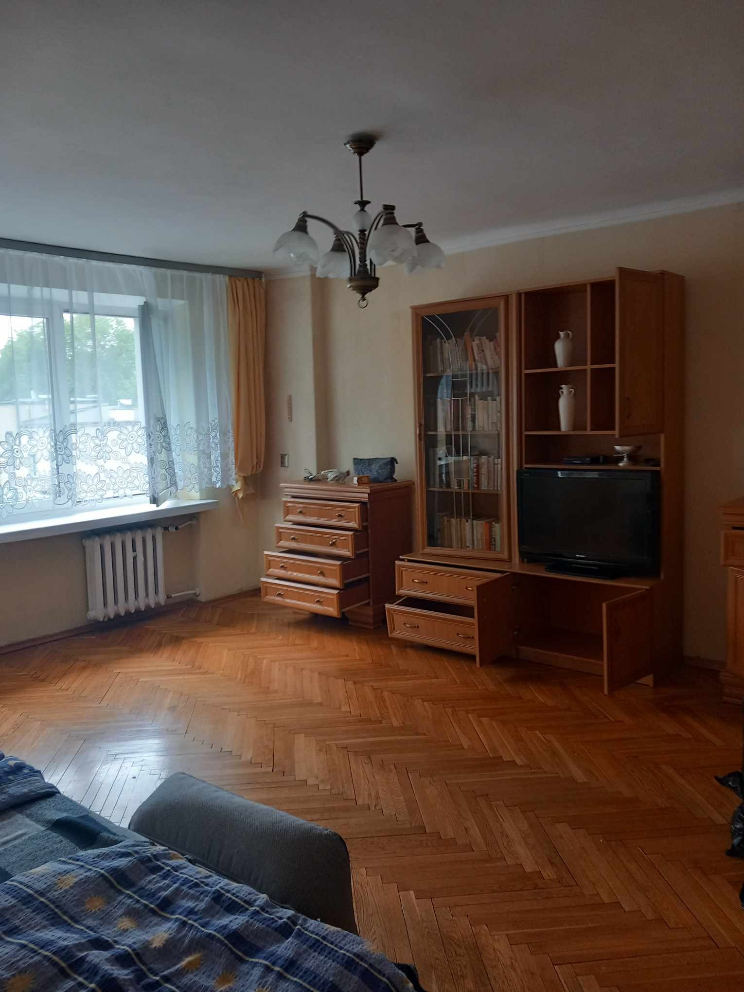 Pokój dzienny z parkietem w jodełkę, drewnianą meblościanką z otwartymi półkami i przeszkloną witryną, oraz komodą z wysuniętymi szufladami, oświetlony przez okno z koronkowymi firankami i żyrandol...