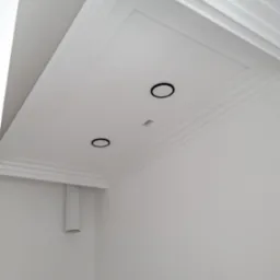 Biały sufit z dekoracyjnymi listwami i dwoma okrągłymi, czarnymi oprawami oświetleniowymi oraz centralnym kwadratowym otworem na oświetlenie, widoczny fragment rury wentylacyjnej przy ścianie.