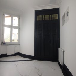 CERAMIKA SYSTEM - Wykończony korytarz z białą marmurową posadzką, czarną stolarką drzwiową i przypodłogową, białe ściany, okno z widokiem na zewnątrz.