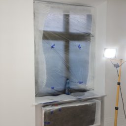 CERAMIKA SYSTEM - Przygotowanie do malowania: okno i grzejnik zabezpieczone folią malarską, butelka wody na parapecie, lampa oświetlająca.
