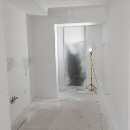 CERAMIKA SYSTEM - Wnętrze pokoju w trakcie malowania na biało, podłoga zabezpieczona folią, widoczny reflektor na statywie, ściany świeżo pomalowane z widocznymi śladami po gładzi, listwy dekoracyjne pod sufitem.