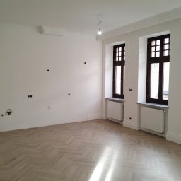 CERAMIKA SYSTEM - Jasny, przestronny pokój po remoncie z parkietem ułożonym we wzór jodełki, białymi ścianami z widocznymi punktami instalacji elektrycznej i trzema oknami z ciemnymi ramami, grzejnikami pod oknami.