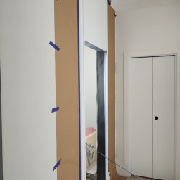 CERAMIKA SYSTEM - Przygotowanie do malowania: ściana i ościeżnica zabezpieczone brązowym papierem i niebieską taśmą malarską, widoczna biała ściana i drzwi w tle.