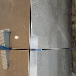 CERAMIKA SYSTEM - Dwie płytki, jedna beżowa, druga szara z marmurkowym wzorem, wyrównane za pomocą klipsów i sznurka poziomującego.