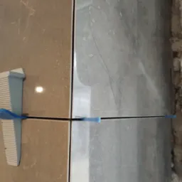 Dwie płytki, jedna beżowa, druga szara z marmurkowym wzorem, wyrównane za pomocą klipsów i sznurka poziomującego.