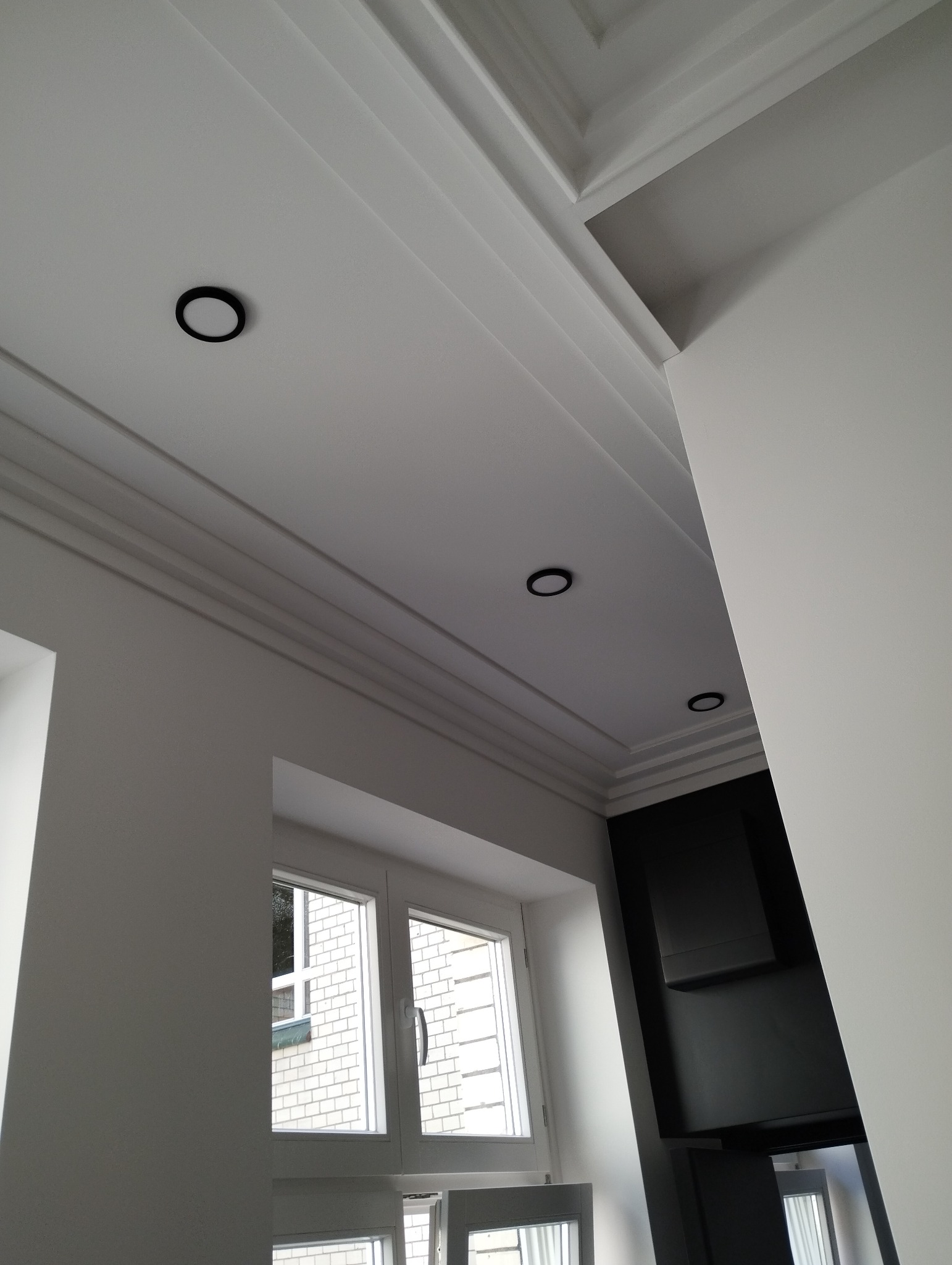 Białe wnętrze z eleganckim sufitem kasetonowym, oświetleniem punktowym w czarnych oprawach, widok na okno i fragment czarnej zabudowy.