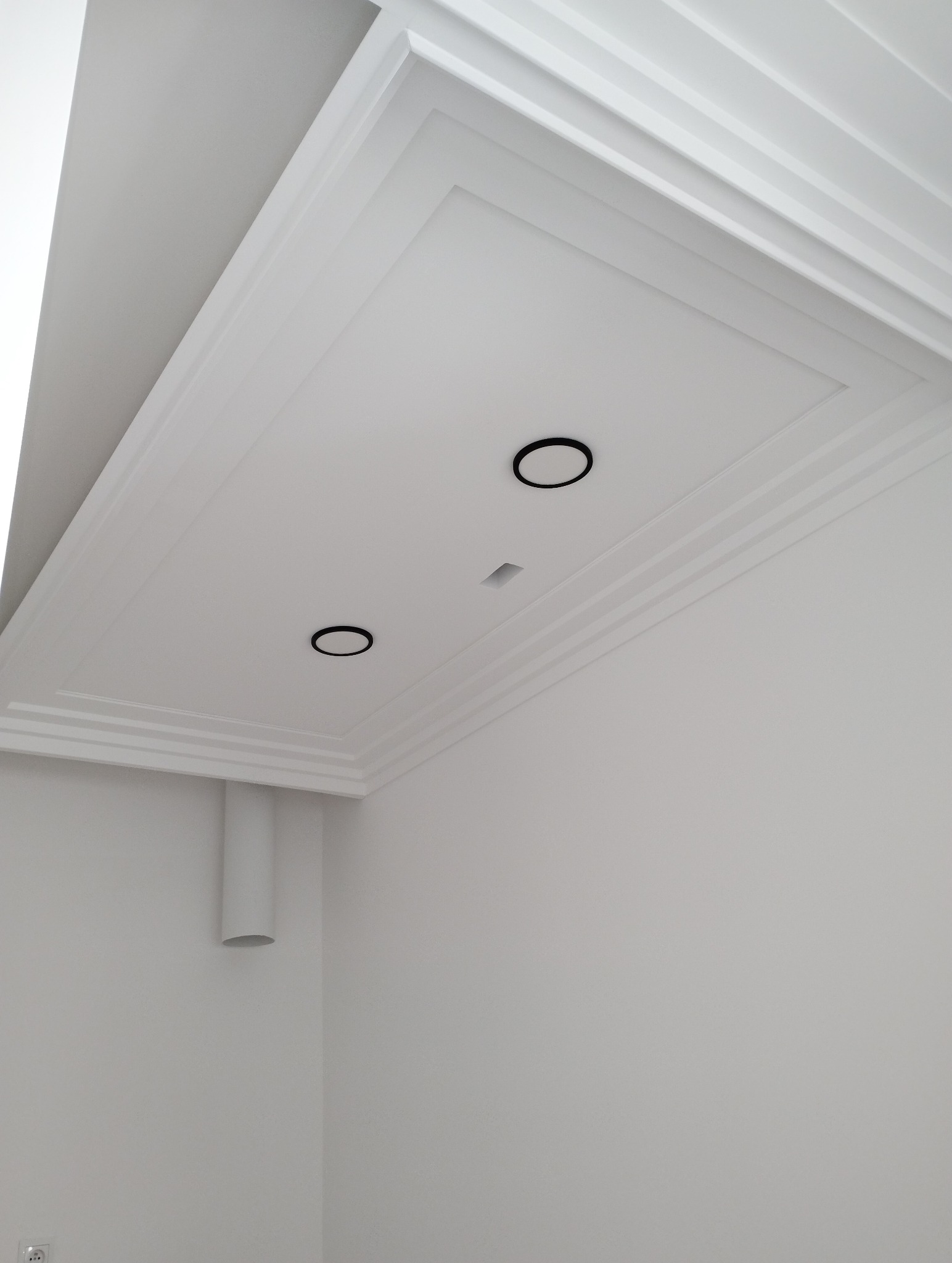 Biały sufit z dekoracyjnymi listwami i dwoma okrągłymi, czarnymi oprawami oświetleniowymi oraz centralnym kwadratowym otworem na oświetlenie, widoczny fragment rury wentylacyjnej przy ścianie.