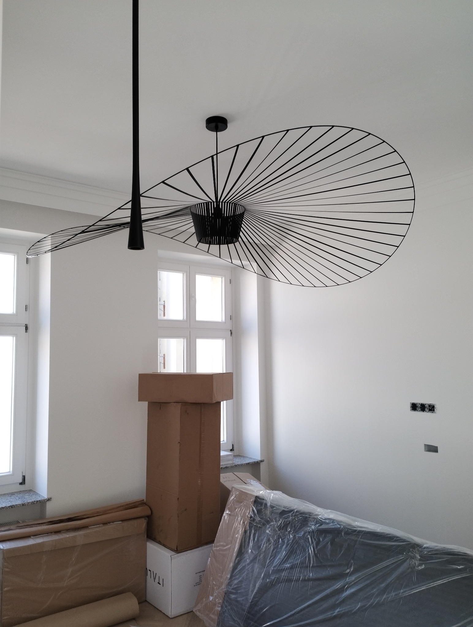 Czarna, designerska lampa sufitowa z metalowymi prętami w kształcie wachlarza, zamontowana w jasnym, odnowionym pokoju z kartonami i meblami zabezpieczonymi folią.
