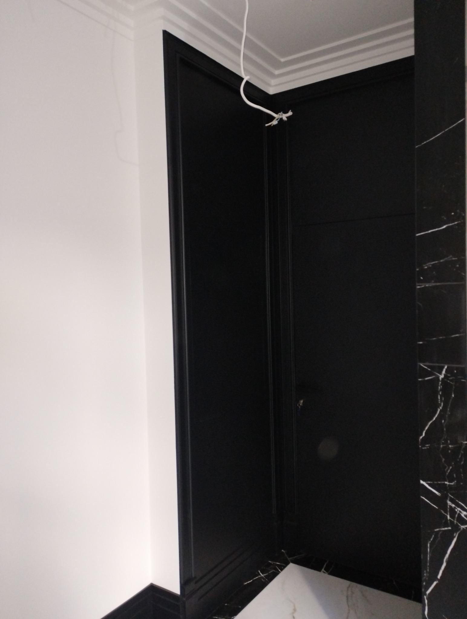 Czarne drzwi wewnętrzne z ościeżnicą w ciemnej tonacji, biała ściana i marmurowe wykończenie fragmentu ściany, widoczne przewody pod sufitem.