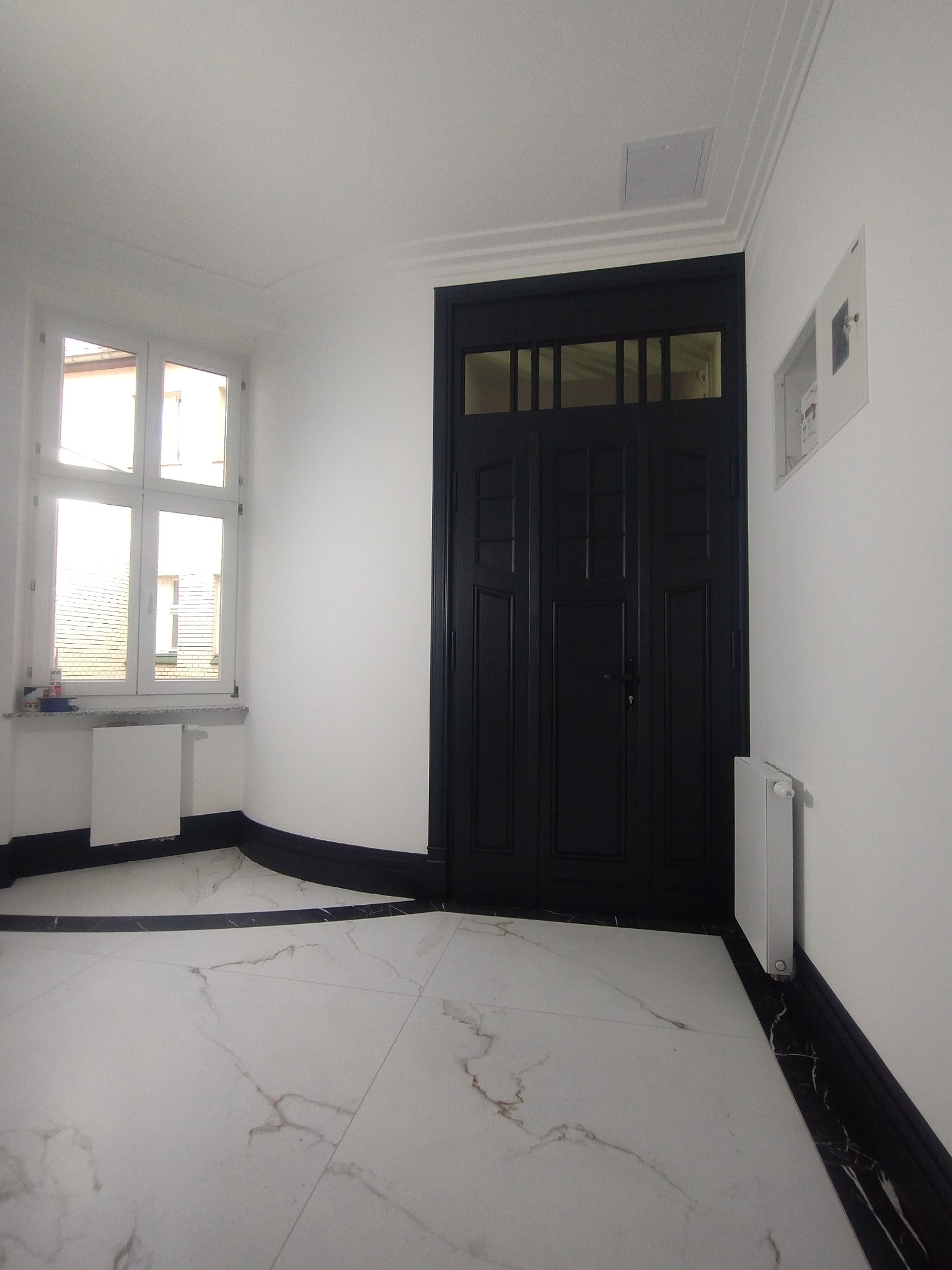 Wykończony korytarz z białą marmurową posadzką, czarną stolarką drzwiową i przypodłogową, białe ściany, okno z widokiem na zewnątrz.