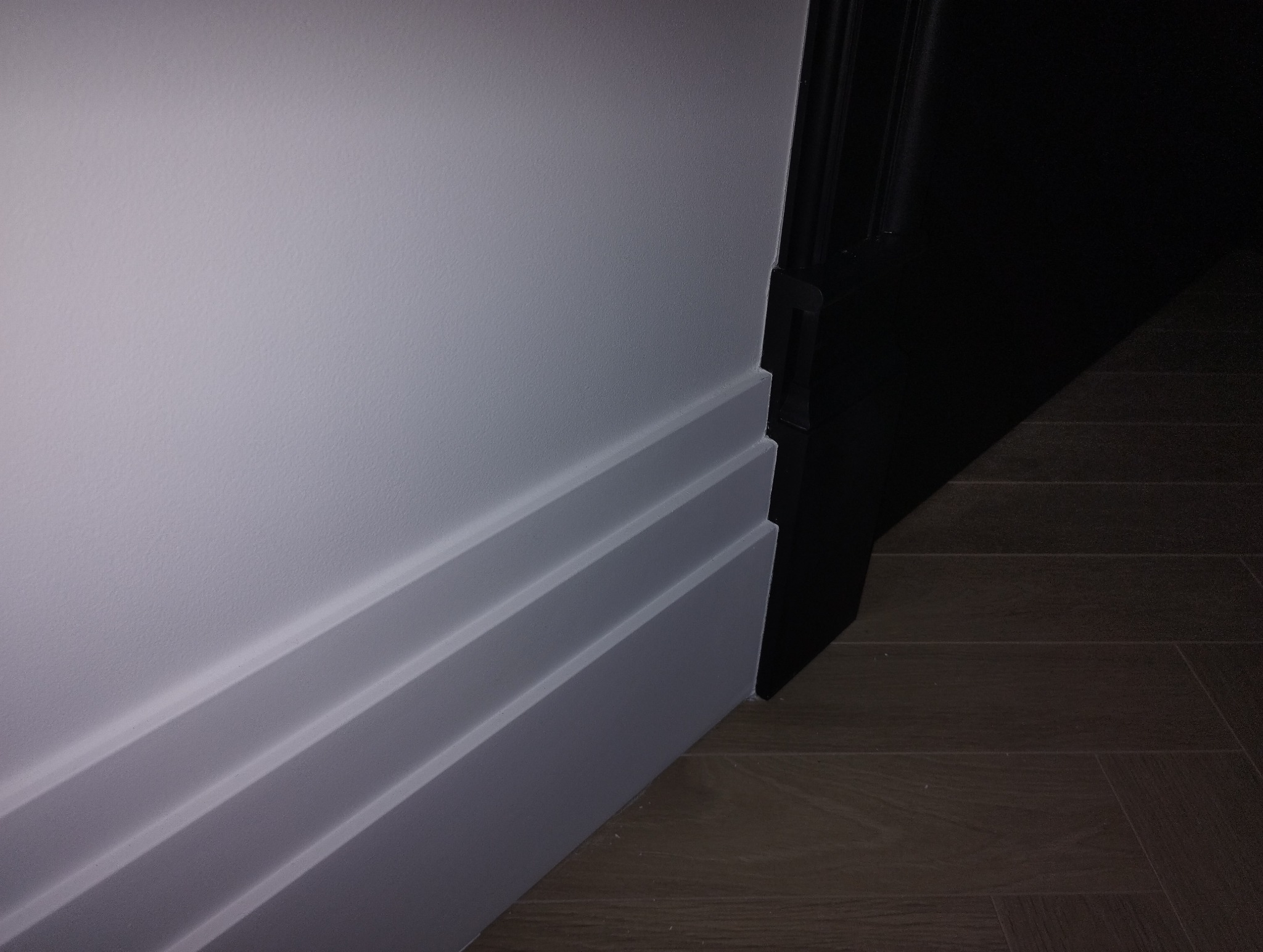 Szara ściana z trzema białymi listwami przypodłogowymi, przy czarnej ościeżnicy drzwiowej i ciemnej podłodze panelowej.