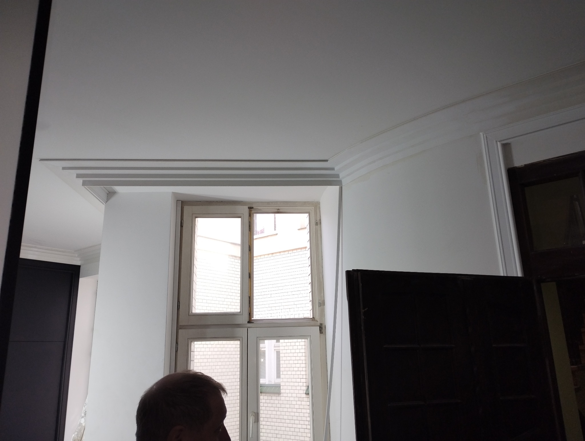 Białe listwy dekoracyjne zamontowane pod sufitem, nad oknem z widokiem na ceglany budynek.