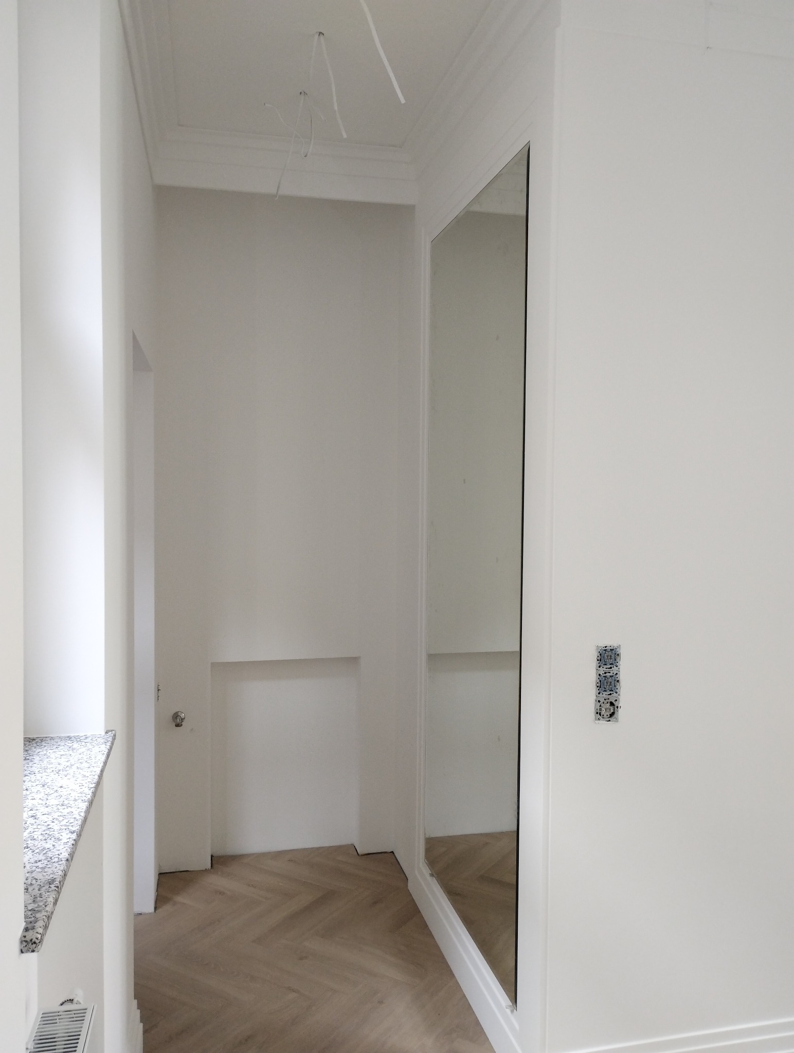 Wnętrze po odświeżeniu: białe ściany, parkiet ułożony w jodełkę, duże lustro w białej ramie, widoczne kable wystające z sufitu, gniazdka elektryczne bez obudowy.