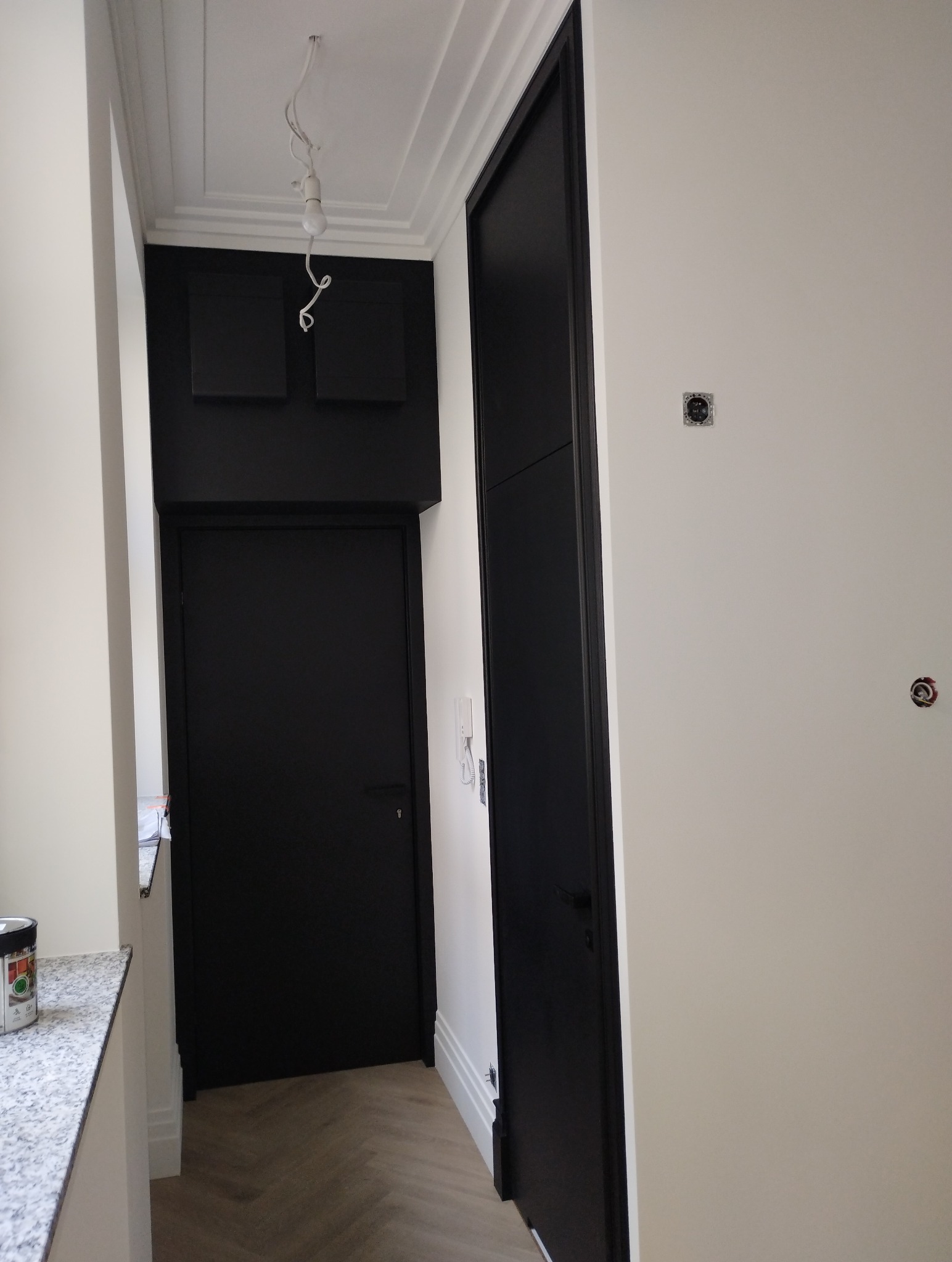 Nowoczesne wnętrze z czarnymi drzwiami i panelami akustycznymi, widoczny fragment podłogi w jodełkę, surowa żarówka zwisająca z sufitu.