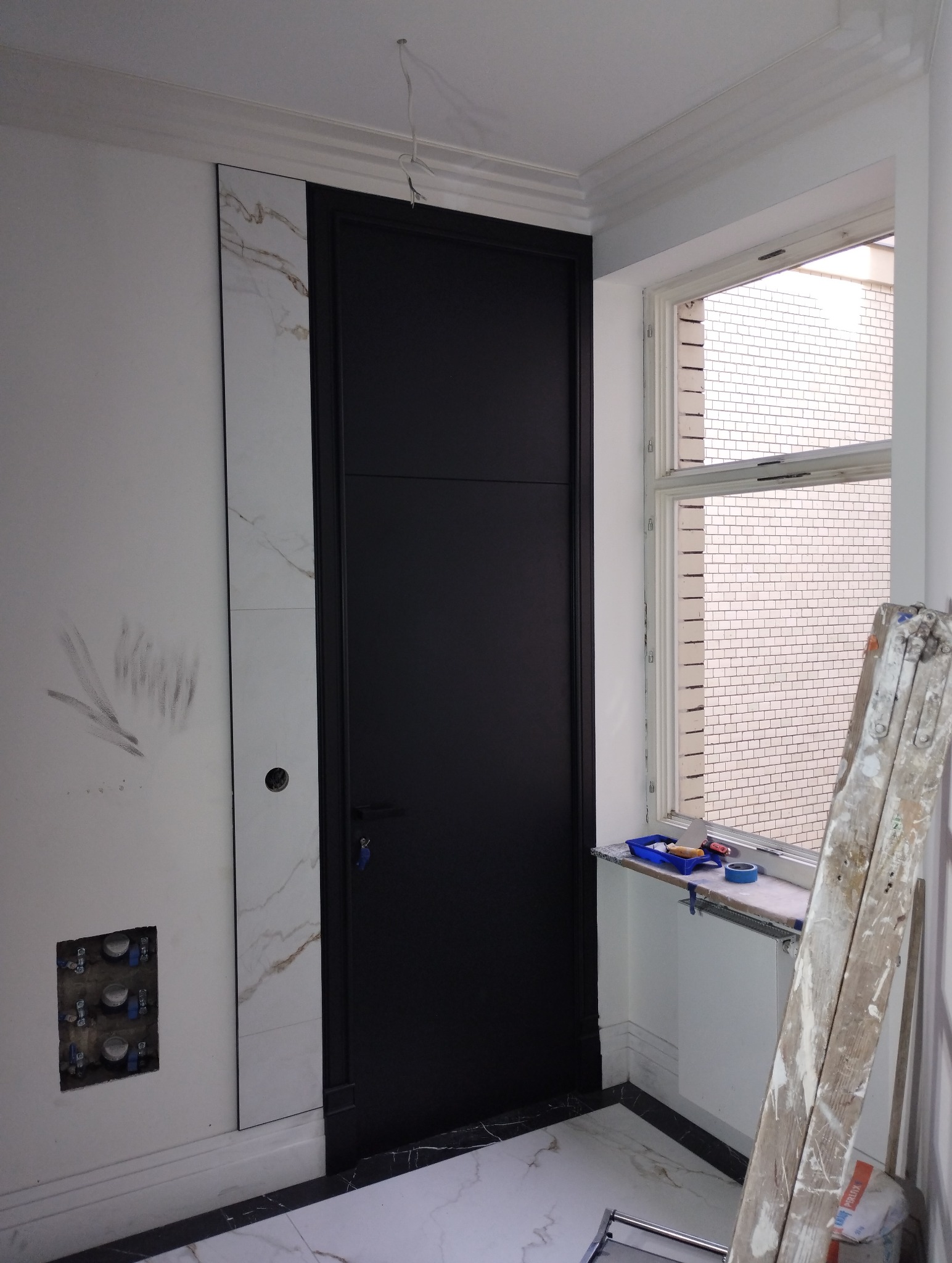 Czarne drzwi wewnętrzne z ościeżnicą, obok fragmentu ściany z płytami imitującymi marmur, widoczne ślady po pracach, drabina oparta o ścianę przy oknie.