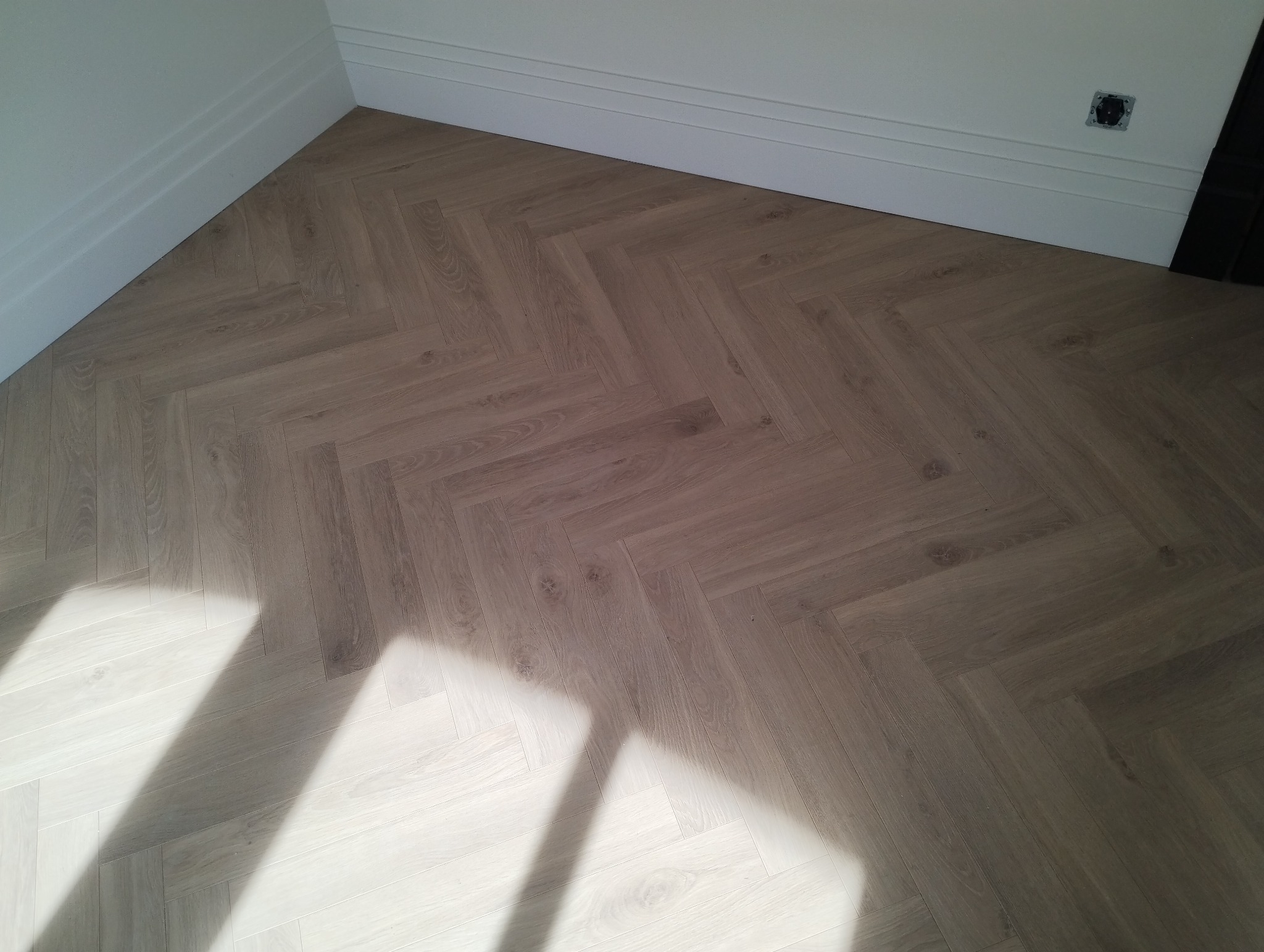 Parkiet ułożony w jodełkę w rogu pokoju w Ostrowie Wielkopolskim, widoczne białe listwy przypodłogowe i cień padający na podłogę.