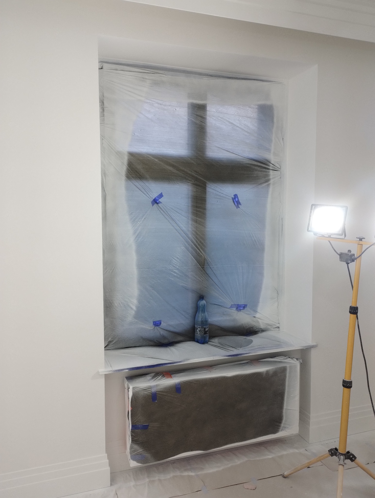 Przygotowanie do malowania: okno i grzejnik zabezpieczone folią malarską, butelka wody na parapecie, lampa oświetlająca.