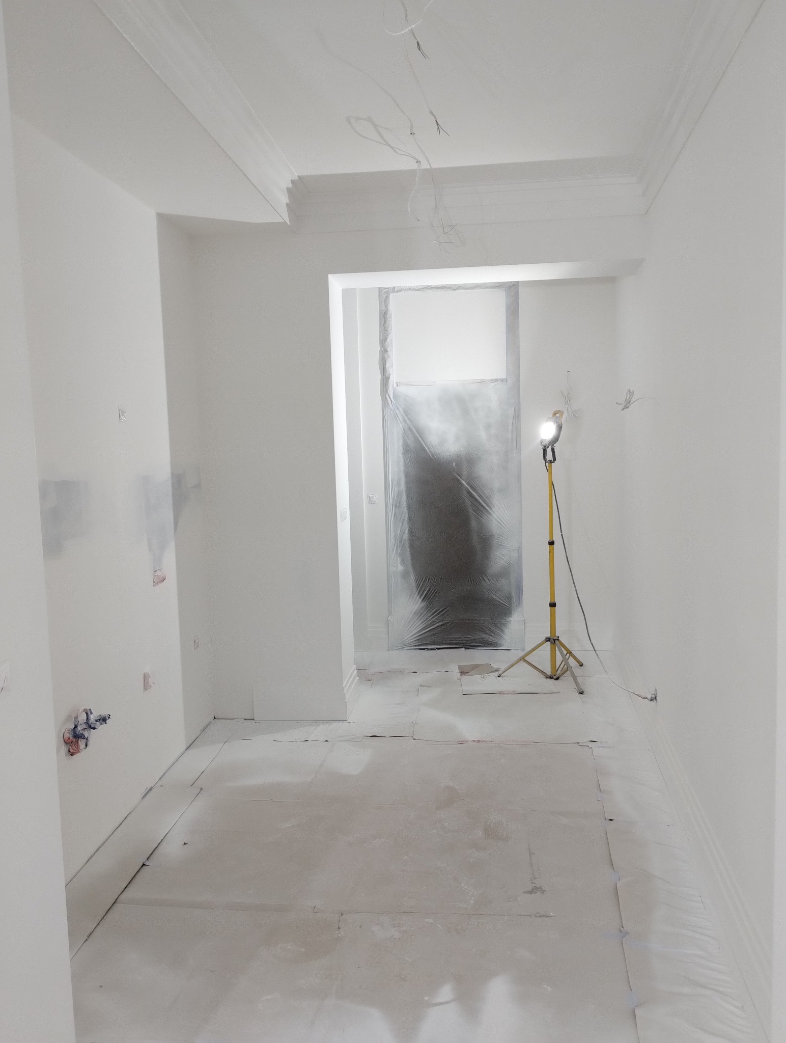 Wnętrze pokoju w trakcie malowania na biało, podłoga zabezpieczona folią, widoczny reflektor na statywie, ściany świeżo pomalowane z widocznymi śladami po gładzi, listwy dekoracyjne pod sufitem.