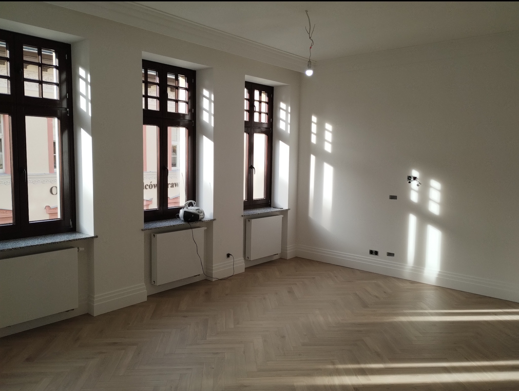 Pusty, jasny pokój z parkietem w jodełkę, trzema oknami z ciemnymi ramami i białymi grzejnikami pod nimi, światło słoneczne tworzy geometryczne wzory na ścianach, widoczne gniazdka elektryczne...