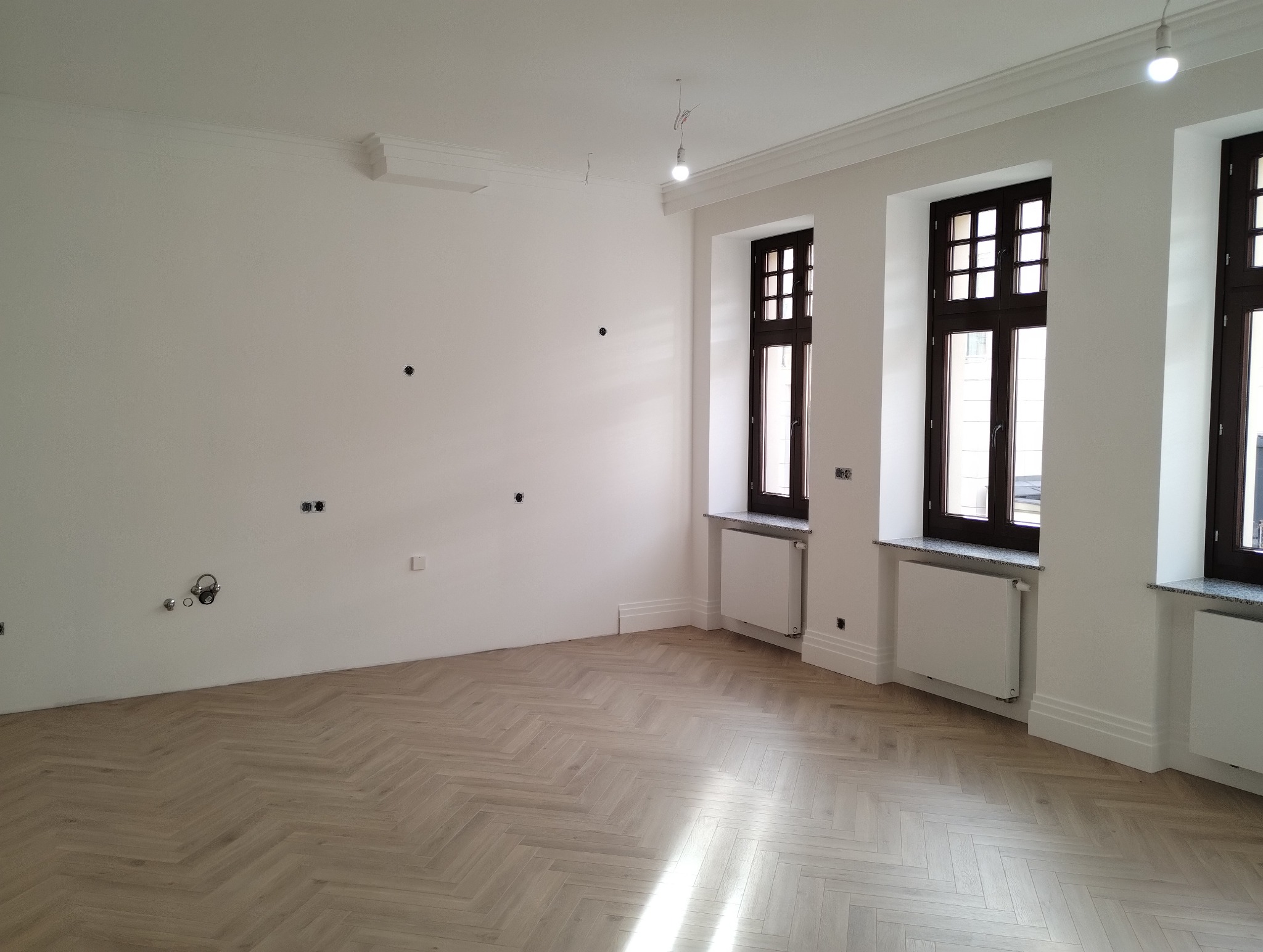 Jasny, przestronny pokój po remoncie z parkietem ułożonym we wzór jodełki, białymi ścianami z widocznymi punktami instalacji elektrycznej i trzema oknami z ciemnymi ramami, grzejnikami pod oknami.