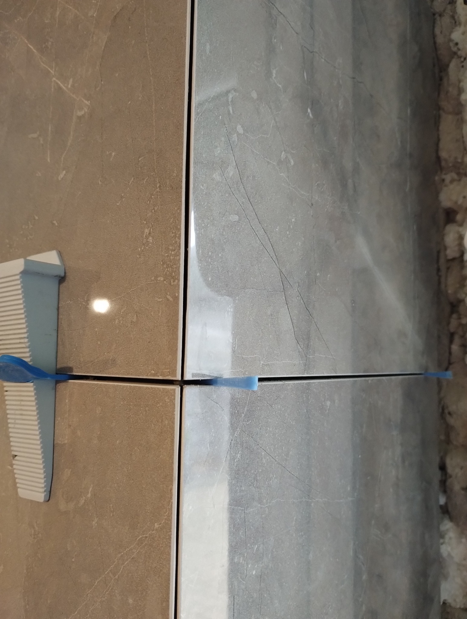 Dwie płytki, jedna beżowa, druga szara z marmurkowym wzorem, wyrównane za pomocą klipsów i sznurka poziomującego.