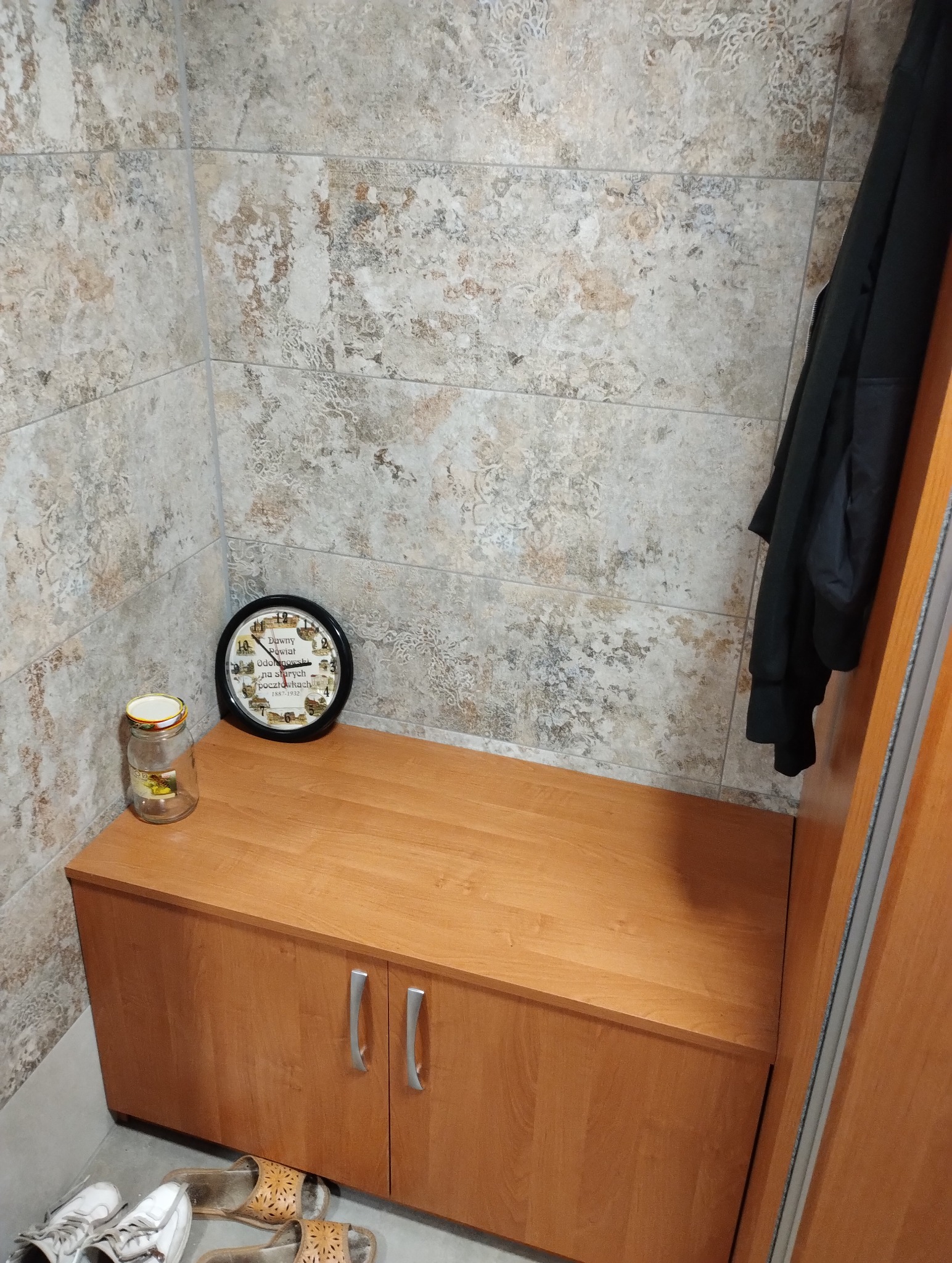 Wykończony fragment przedpokoju z beżowymi płytkami imitującymi kamień, drewnianą szafką z dwoma drzwiczkami, zegarem z motywami retro i czarną kurtką wiszącą na ścianie.