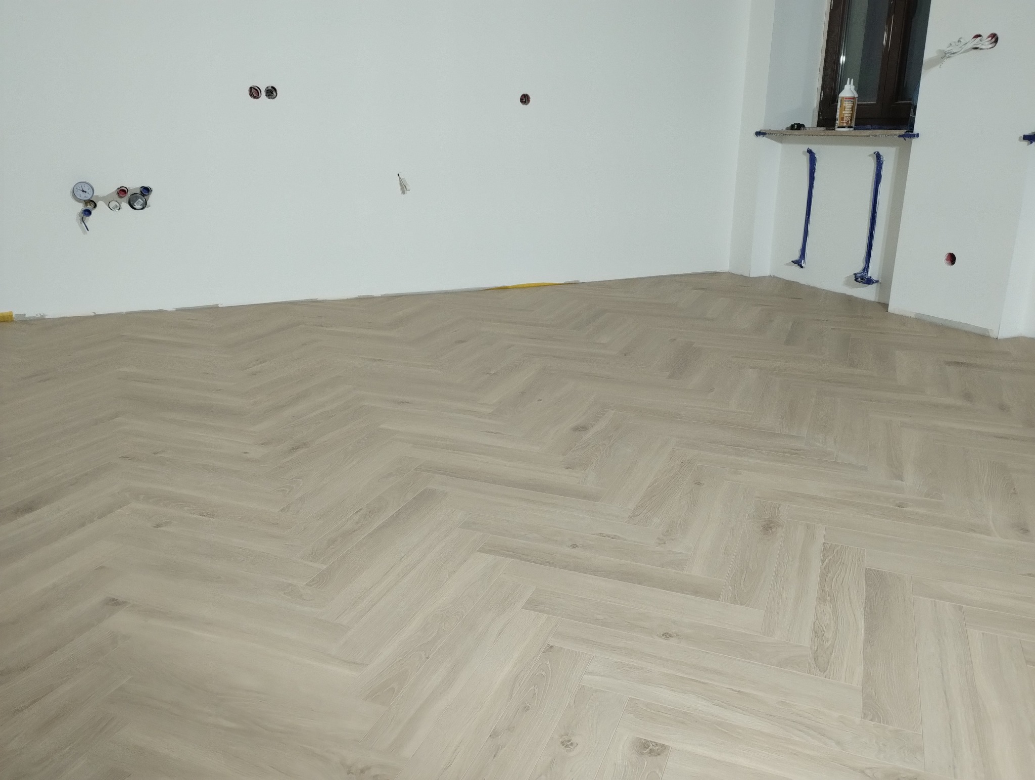 Jasne panele ułożone we wzór jodełki w pokoju z białymi ścianami i instalacjami hydraulicznymi.