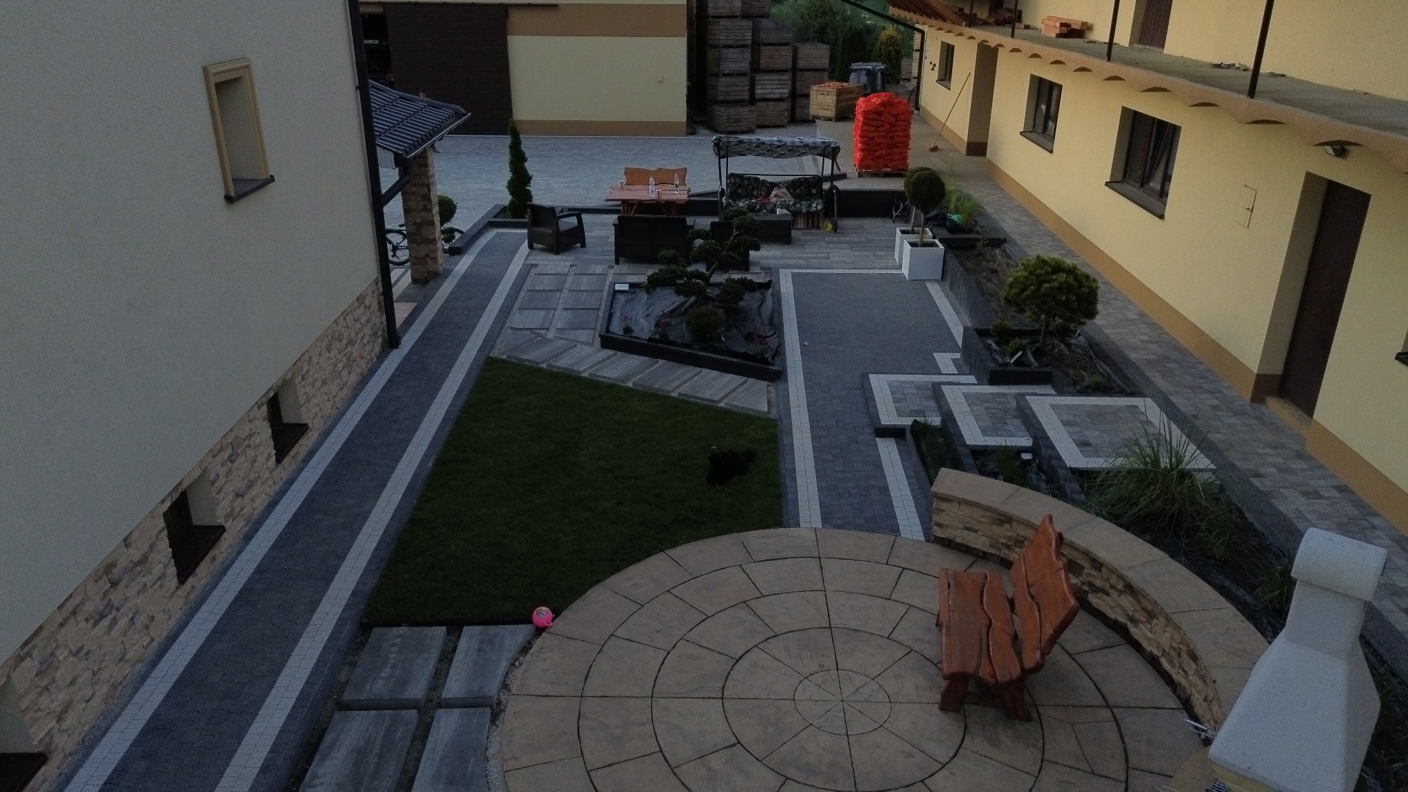 Widok z góry na starannie zaprojektowany ogród z kostki brukowej, trawnika i roślin ozdobnych. Okrągły taras z ławką, ścieżki z kostki, elementy małej architektury.