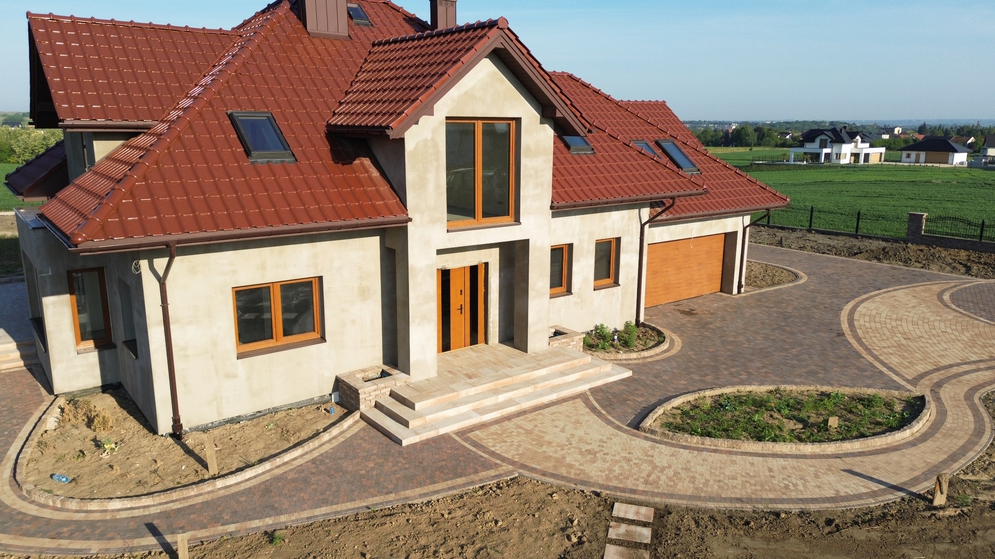 Nowoczesny dom z brązowym dachem i kostką brukową ułożoną w dekoracyjny wzór na podjeździe. Widok z góry na posesję w słoneczny dzień.