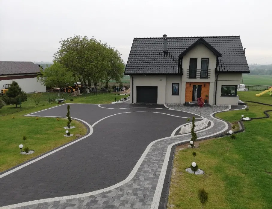 Nowoczesny dom z eleganckim podjazdem z ciemnej kostki brukowej, obrzeża z jasnej kostki. Zadbany trawnik i oświetlenie ogrodowe podkreślają estetykę posesji.