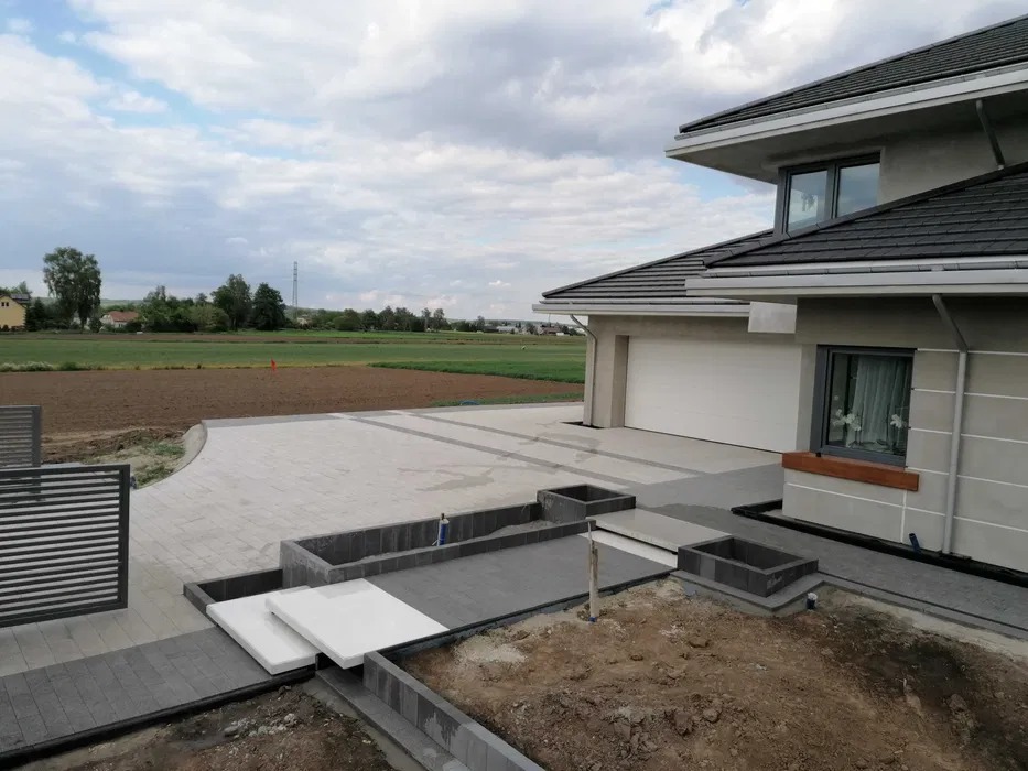 Nowoczesny podjazd z kostki brukowej w odcieniach szarości prowadzący do garażu. Widoczne elementy dekoracyjne i rabaty kwiatowe w trakcie realizacji. Tło stanowi krajobraz polny.