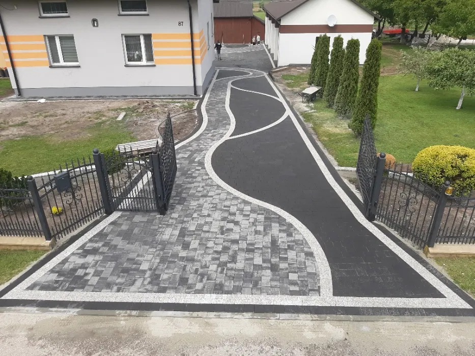 Ułożona kostka brukowa w dwóch odcieniach szarości tworząca wzór ścieżki przed domem z otwartą, ozdobną bramą. Widok z góry.