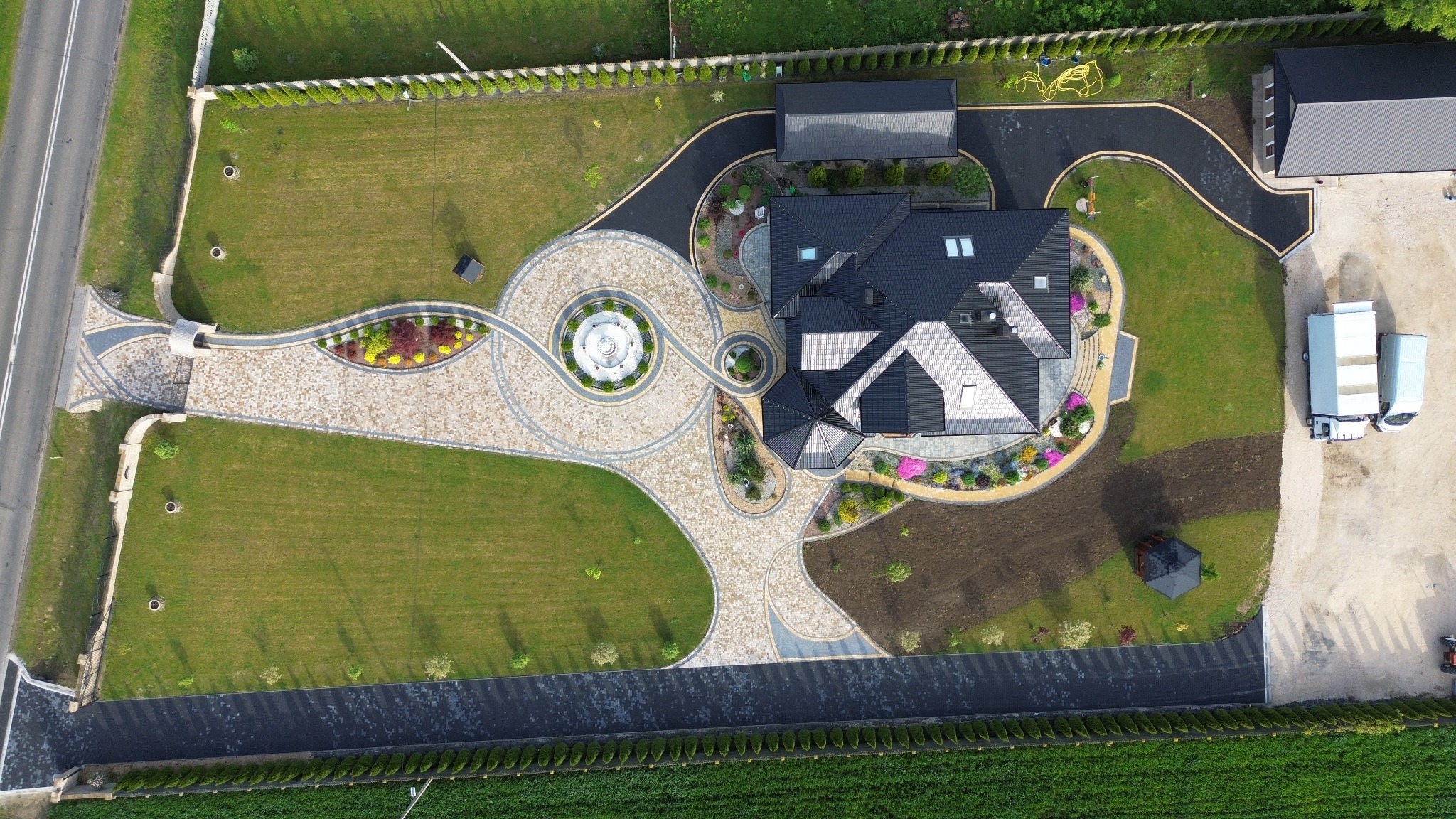 Widok z lotu ptaka na posesję z fontanną, ozdobnymi ścieżkami z kostki brukowej, zadbanym trawnikiem i rabatami kwiatowymi. Asfaltowa droga dojazdowa.