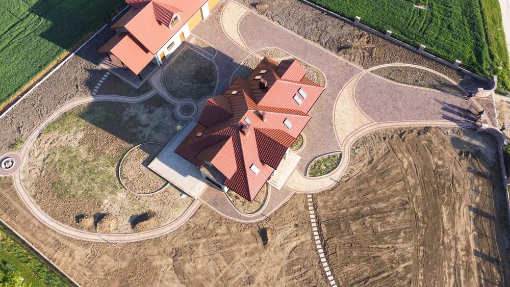 Widok z góry na dom z czerwoną dachówką i ozdobną kostką brukową na podjeździe, otoczony przygotowanym terenem pod trawnik i zieleń. Częściowo widoczny drugi budynek.