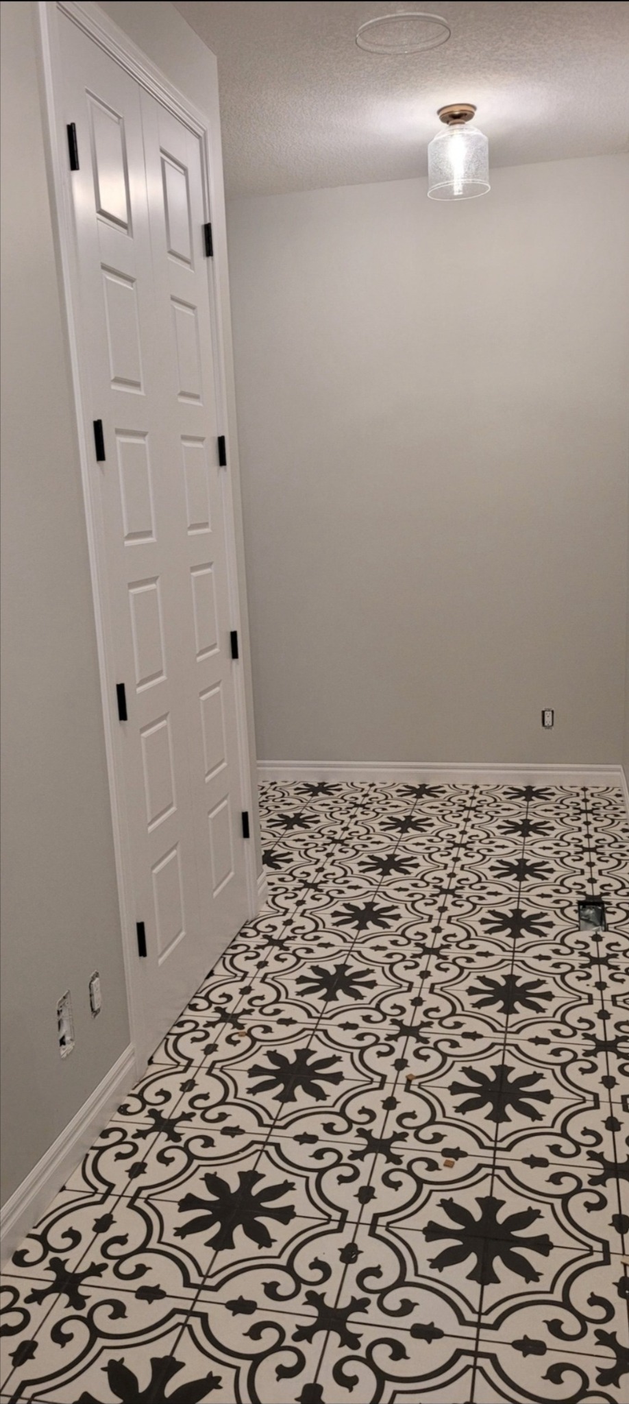 Wnętrze z nową, wzorzystą czarno-białą podłogą z płytek, białe ściany i drzwi z czarnymi klamkami, widoczne niedokończone gniazdka elektryczne.