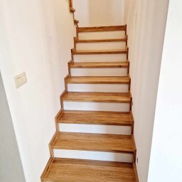 Mateusz Górski - Dębowe schody z litego drewna, zamontowane na betonowej konstrukcji, widoczne z dołu, z białymi podstopnicami i drewnianą listwą przyścienną.