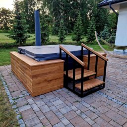 Mateusz Górski - Obudowa jacuzzi z drewna z wbudowanymi szafkami i schodkami z metalową ramą, na zewnątrz, obok domu z hamakiem.