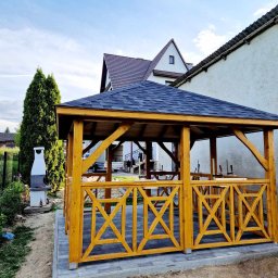 Mateusz Górski - Żółta, drewniana altanka ogrodowa z dekoracyjnymi panelami w kształcie litery X, na wybrukowanym tarasie, z grillem obok i domami w tle.