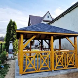 Żółta, drewniana altanka ogrodowa z dekoracyjnymi panelami w kształcie litery X, na wybrukowanym tarasie, z grillem obok i domami w tle.
