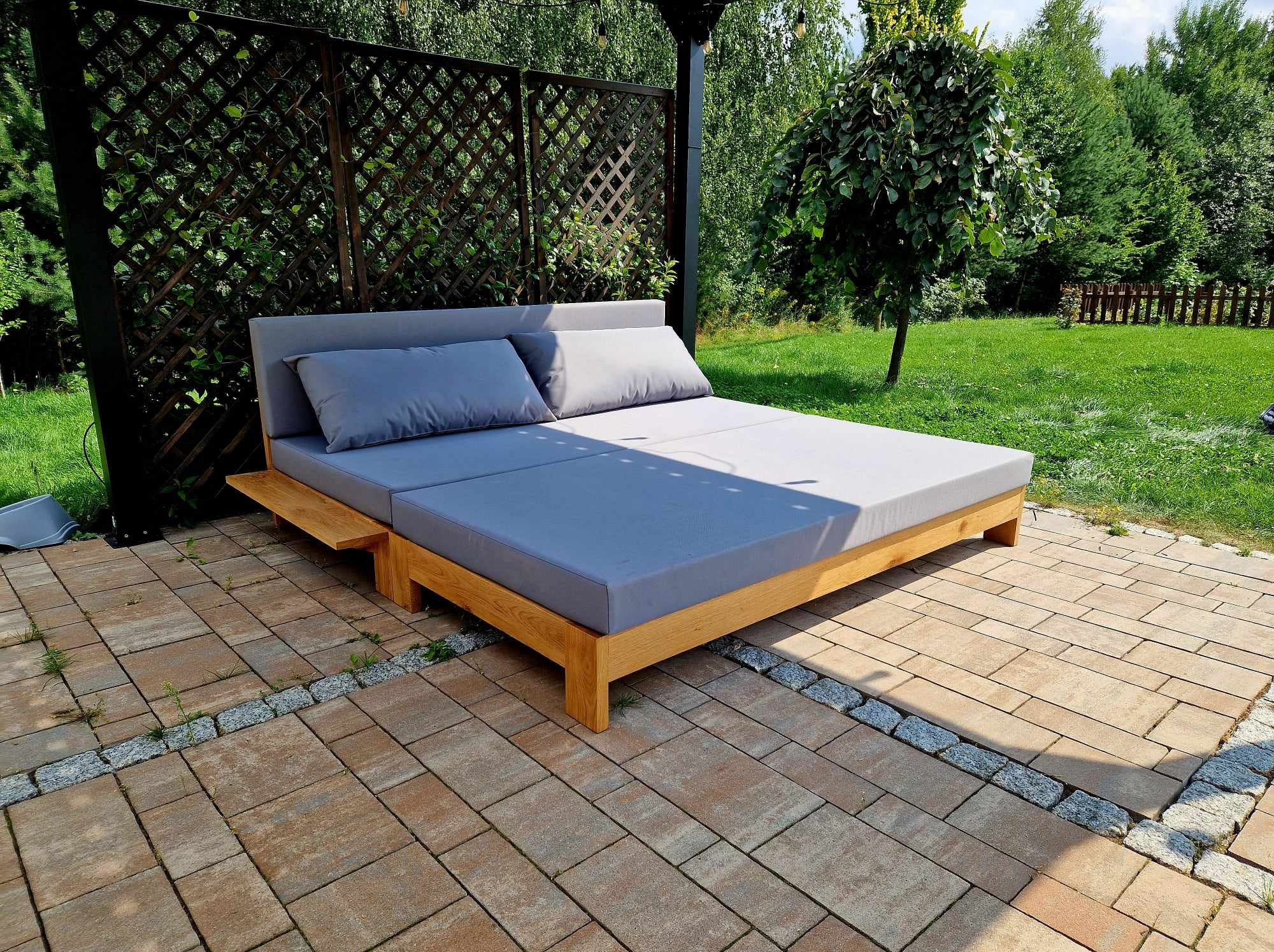 Drewniane łóżko ogrodowe z szarą tapicerką i poduszkami, umieszczone na tarasie z kostki brukowej, w tle zielony trawnik i dekoracyjna drewniana pergola.