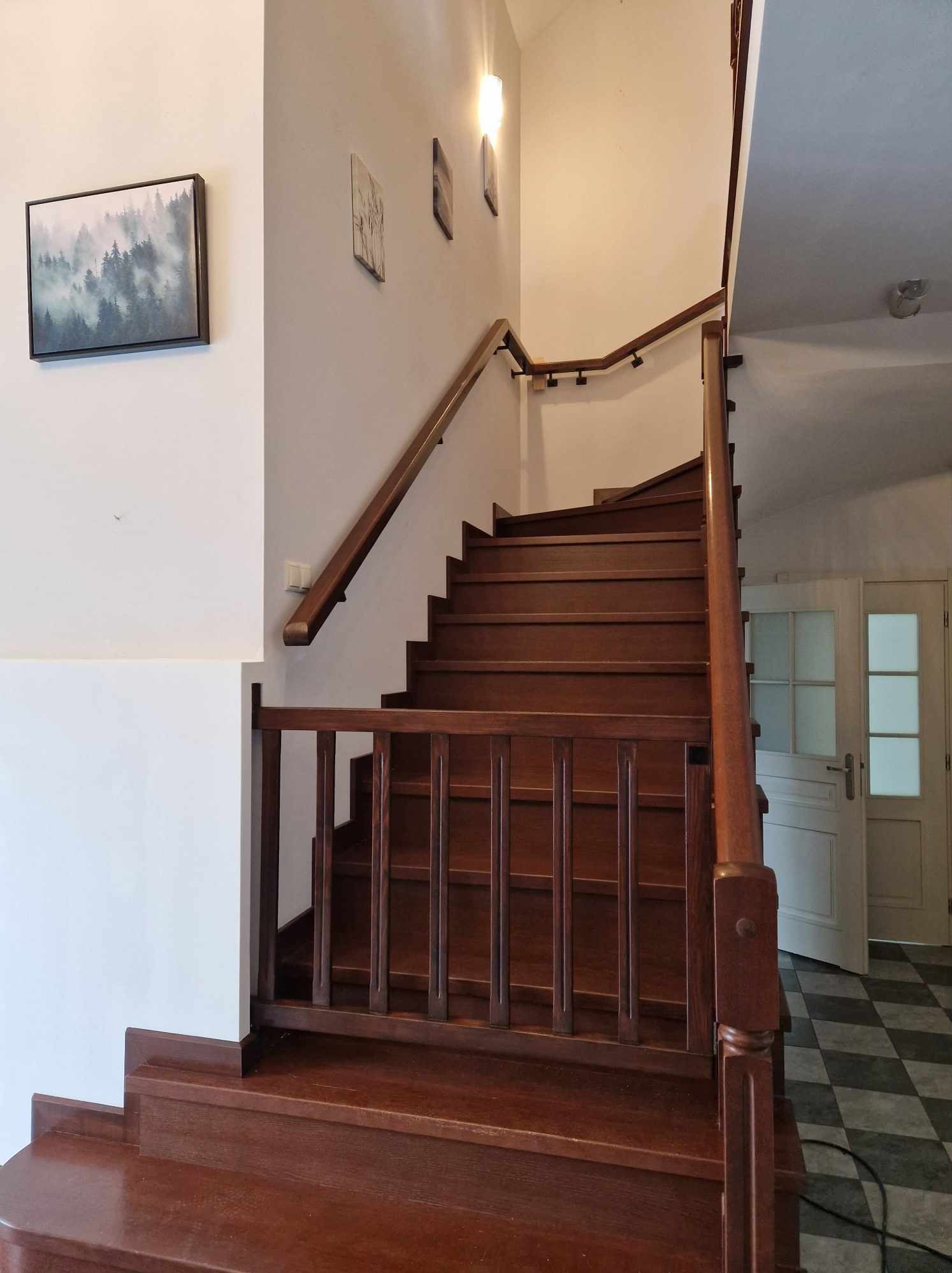 Klasyczne, ciemne stopnie z drewna prowadzące na górę, z drewnianą balustradą i poręczą, na ścianie wiszą obrazy, widoczna podłoga w szachownicę i drzwi.