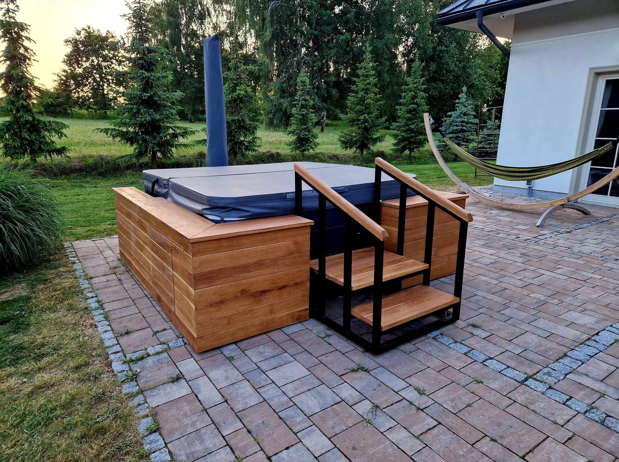Obudowa jacuzzi z drewna z wbudowanymi szafkami i schodkami z metalową ramą, na zewnątrz, obok domu z hamakiem.