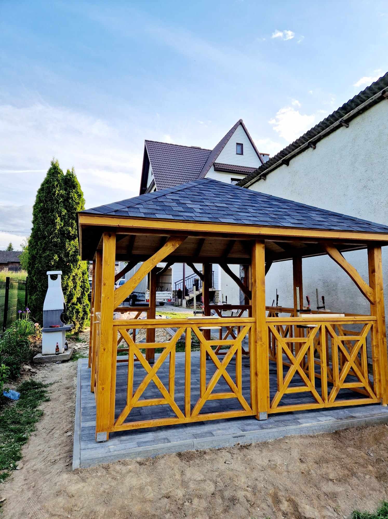 Żółta, drewniana altanka ogrodowa z dekoracyjnymi panelami w kształcie litery X, na wybrukowanym tarasie, z grillem obok i domami w tle.