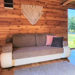 Wnętrze drewnianego domku letniskowego z kanapą, makramą na ścianie i widokiem na zieleń za oknem.