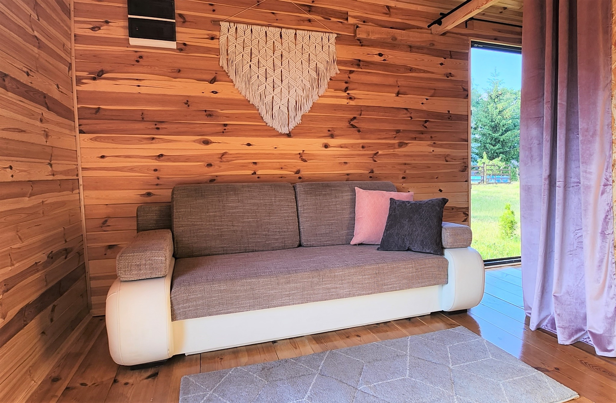 Wnętrze drewnianego domku letniskowego z kanapą, makramą na ścianie i widokiem na zieleń za oknem.
