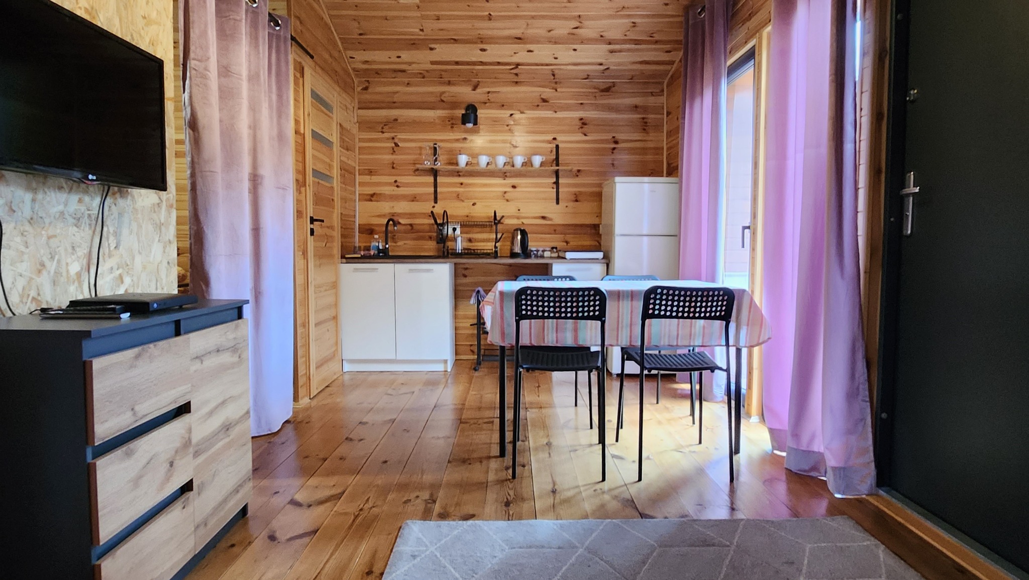 Wnętrze drewnianego domku letniskowego z aneksem kuchennym, stołem z krzesłami i telewizorem, zasłony w odcieniach fioletu.