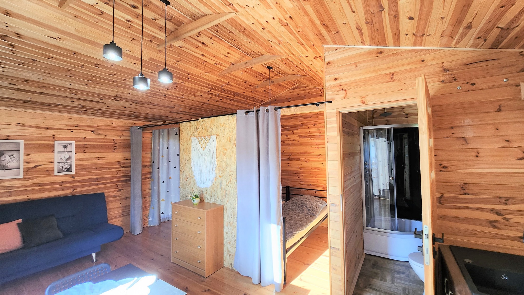 Wnętrze drewnianego domku letniskowego z sypialnią oddzieloną zasłonami, widoczna sofa, komoda z kwiatami, łazienka z kabiną prysznicową i drewniane panele na ścianach i suficie.